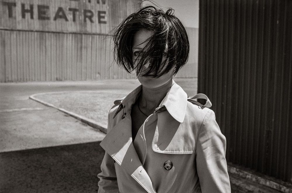 Black and white photo of a person in a trench coat with wind-blown hair covering their face near a theatre.