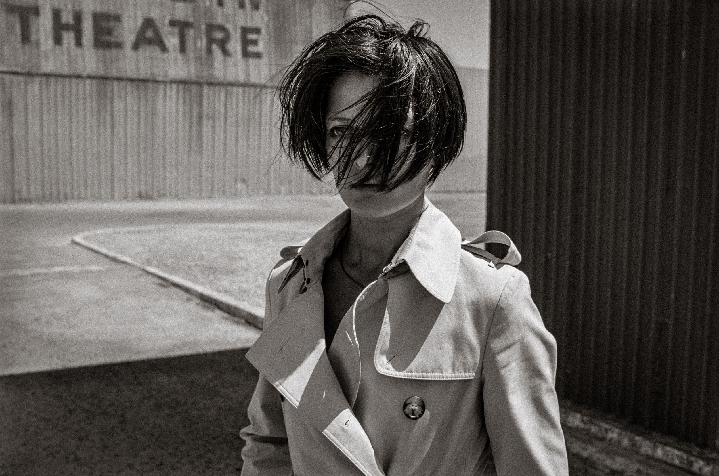 Black and white photo of a person in a trench coat with wind-blown hair covering their face near a theatre.