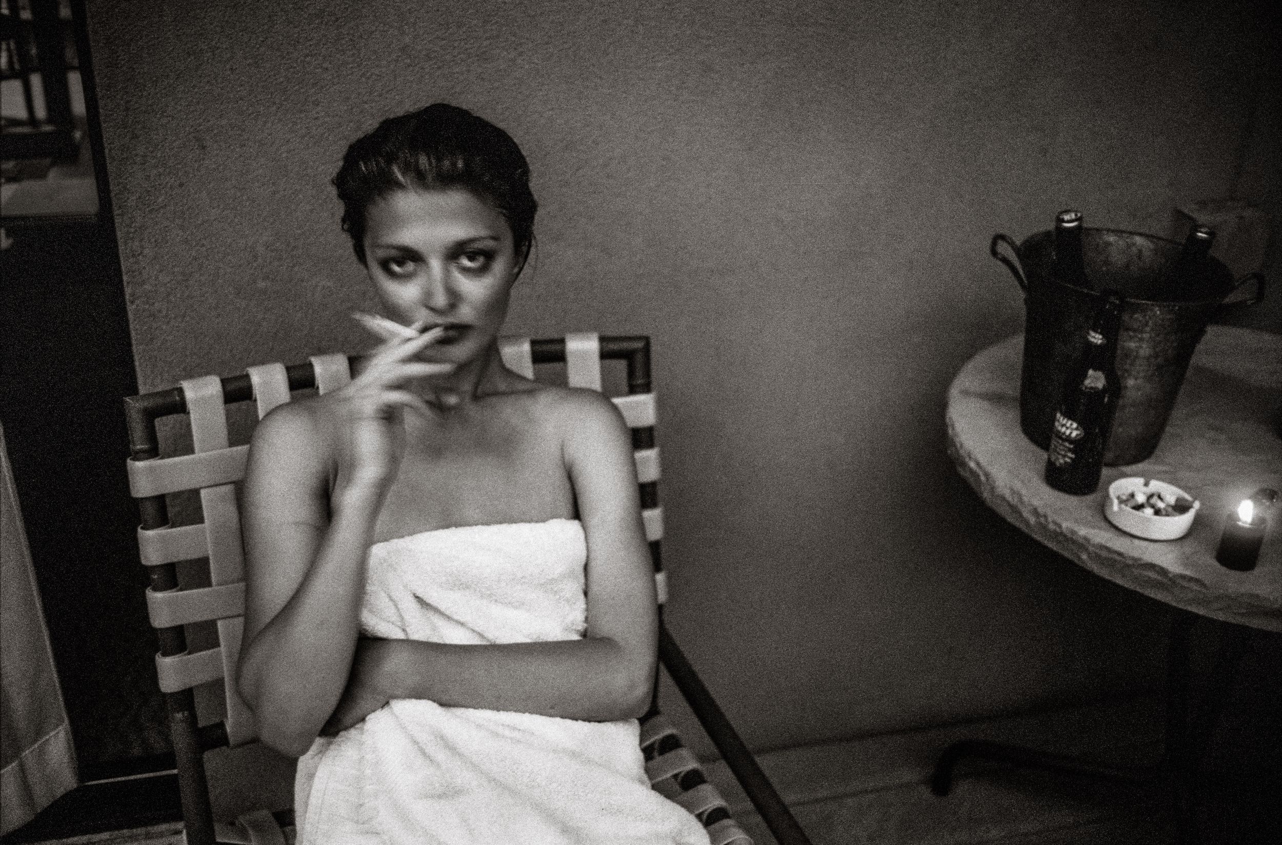 Black and white photo of a young woman wrapped in a towel, sitting in a chair and smoking a cigarette indoors.