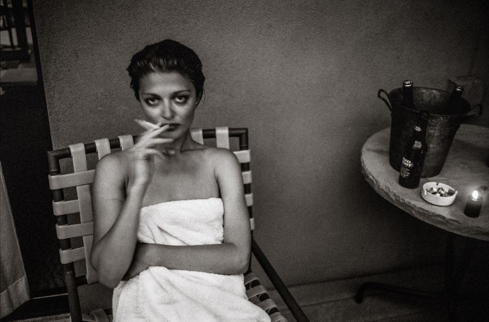 Black and white photo of a young woman wrapped in a towel, sitting in a chair and smoking a cigarette indoors.