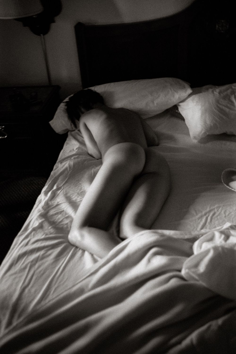Black and white photo of a nude person lying curled on white bed sheets in soft dramatic lighting.
