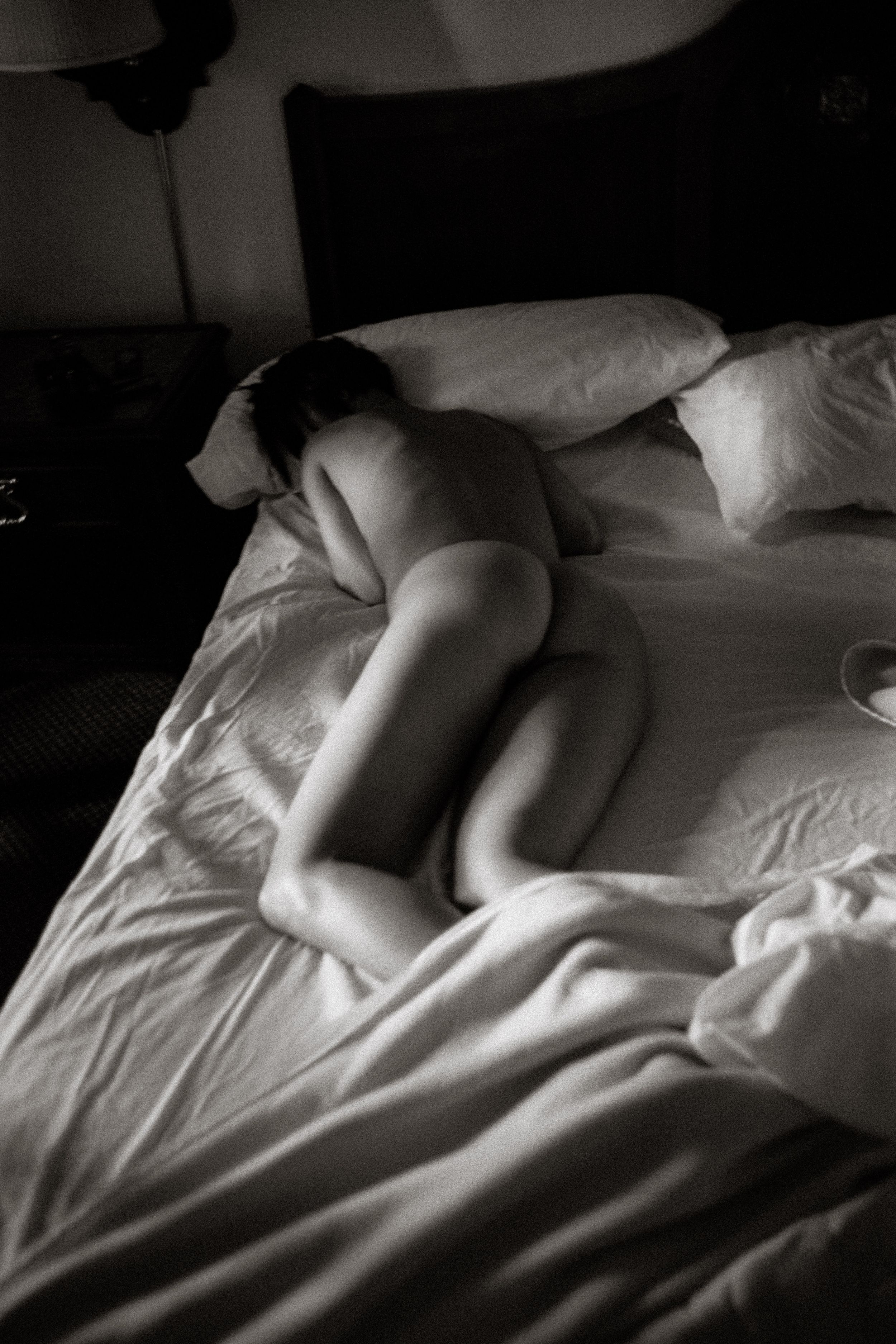 Black and white photo of a nude person lying curled on white bed sheets in soft dramatic lighting.