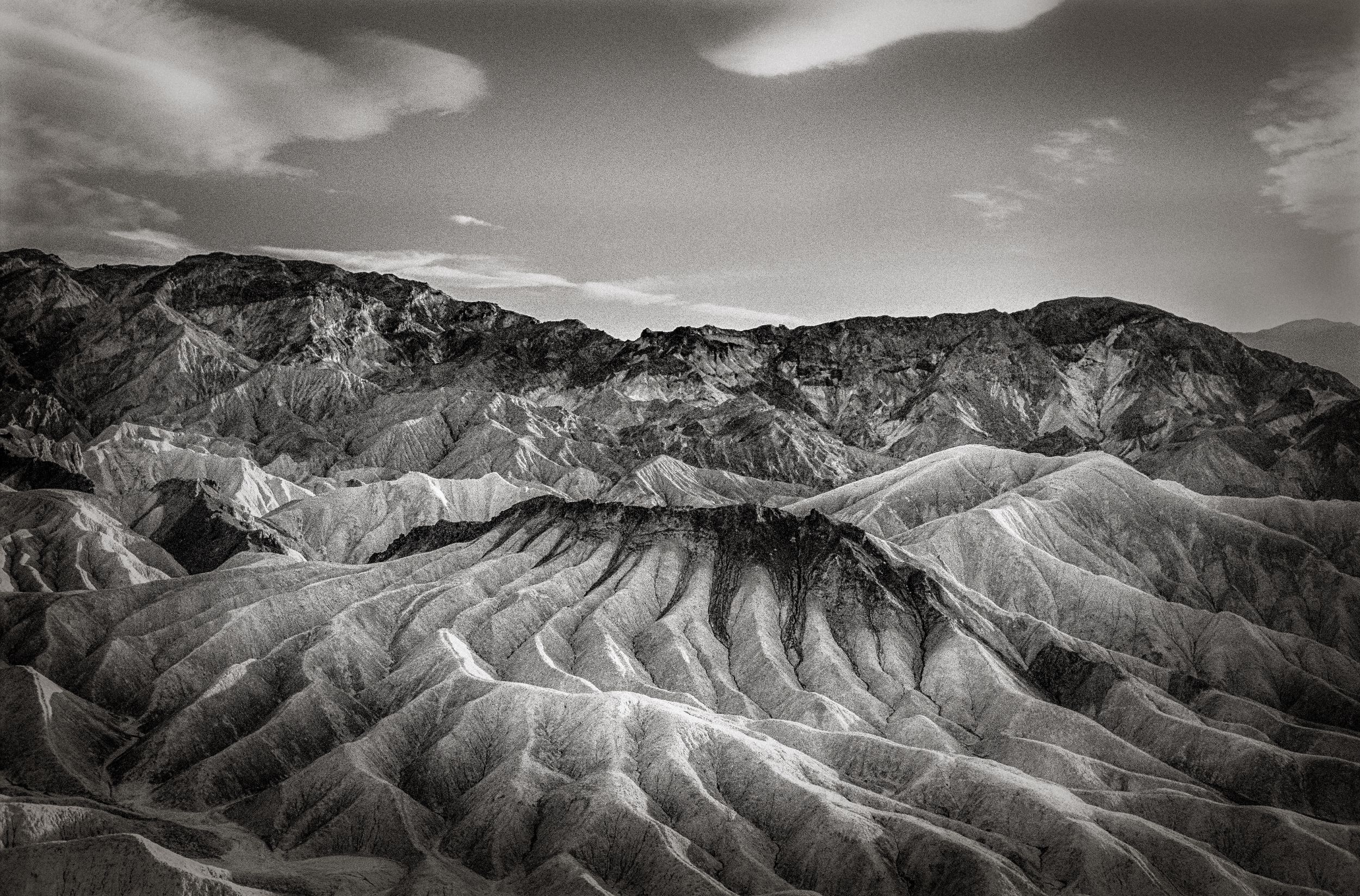 Black and white aerial view of dramatic eroded badlands terrain with layered rock formations under cloudy sky.
