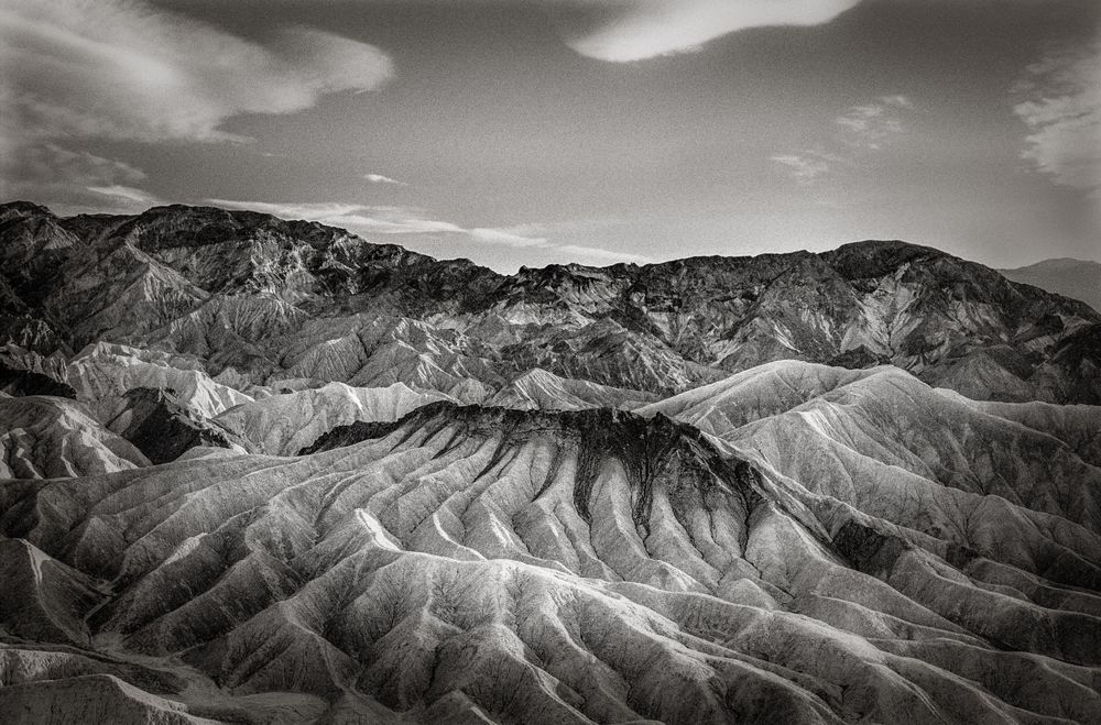 Black and white aerial view of dramatic eroded badlands terrain with layered rock formations under cloudy sky.