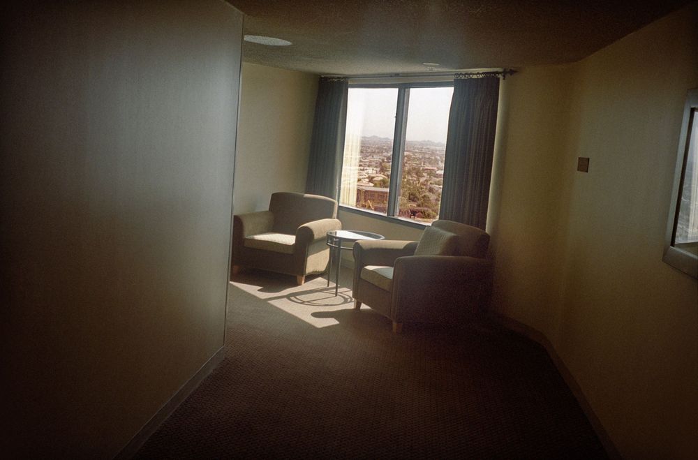 Minimalist hotel room with two armchairs facing a large window overlooking a city skyline.