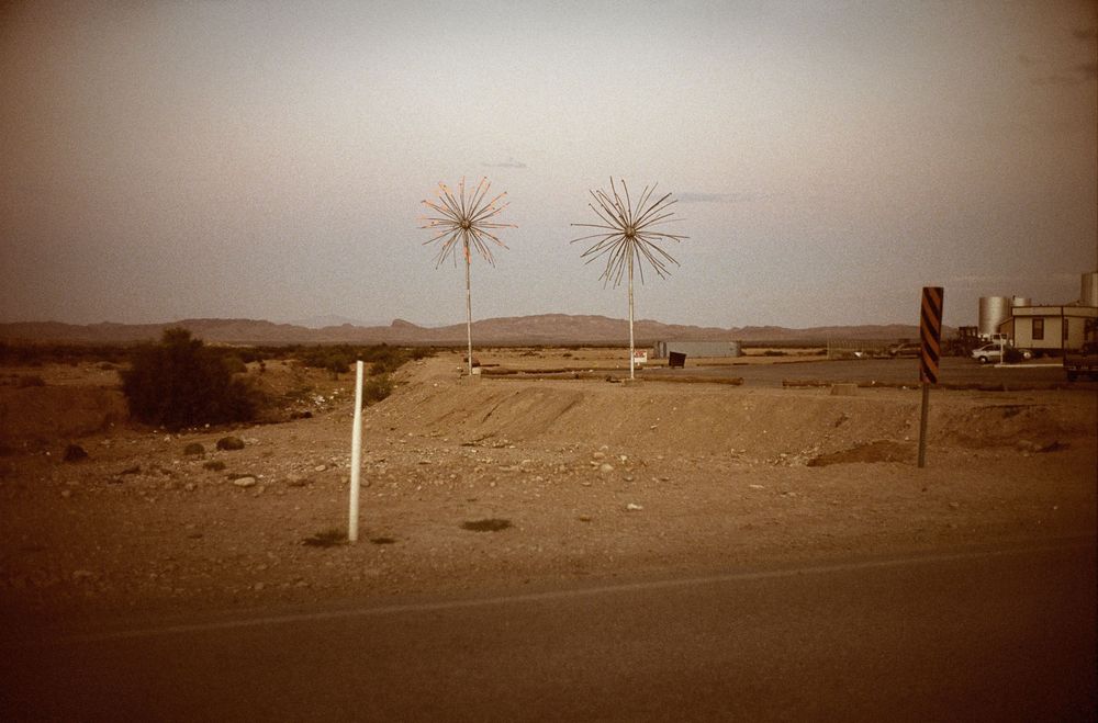 Two starburst metal sculptures stand in a barren desert landscape along a dusty roadside under a hazy sky.