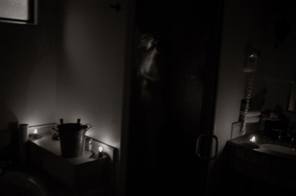 Dimly lit room with candles glowing on surfaces and a figure standing in a doorway in dark atmospheric black and white photo.