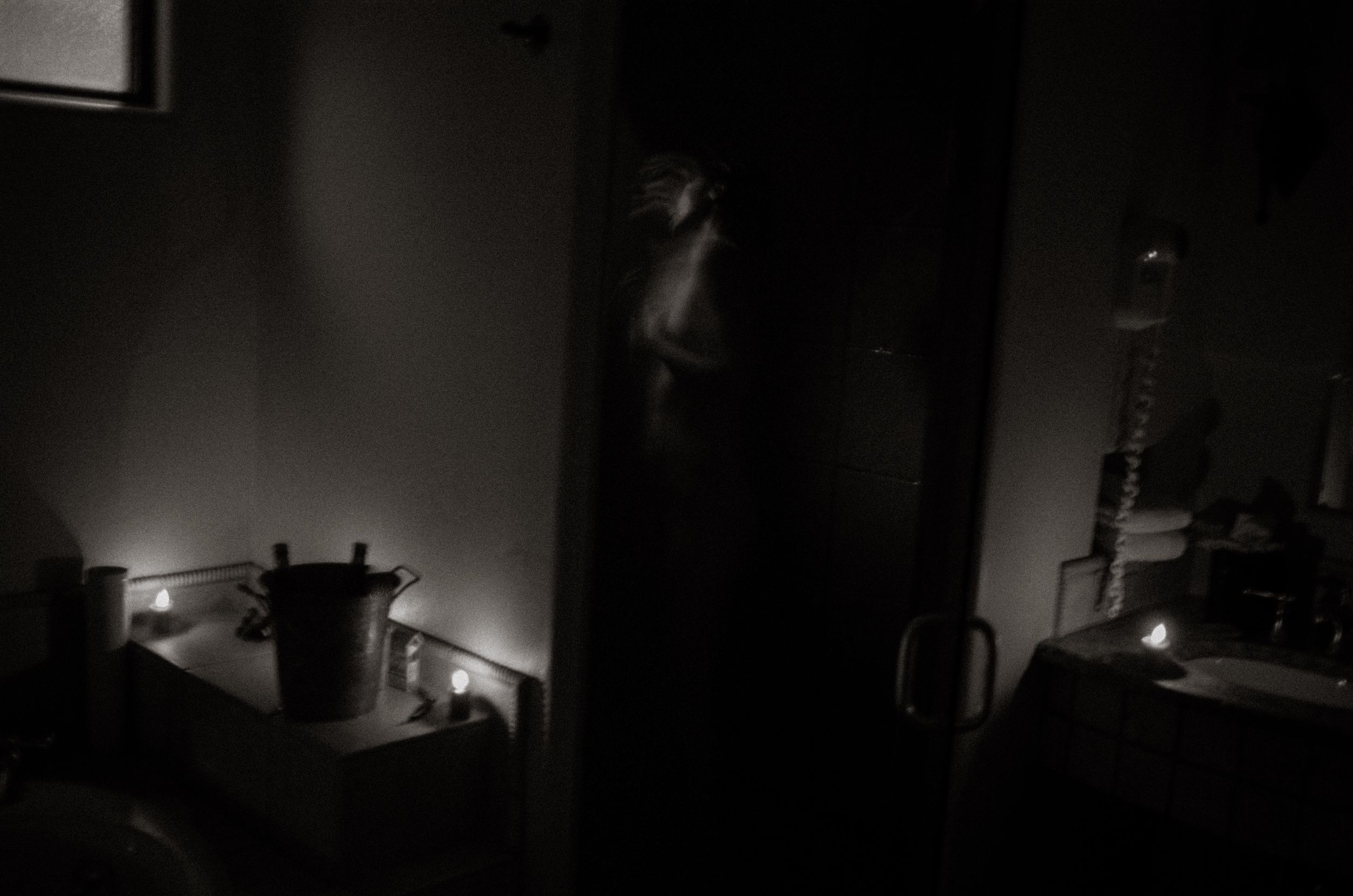 Dimly lit room with candles glowing on surfaces and a figure standing in a doorway in dark atmospheric black and white photo.