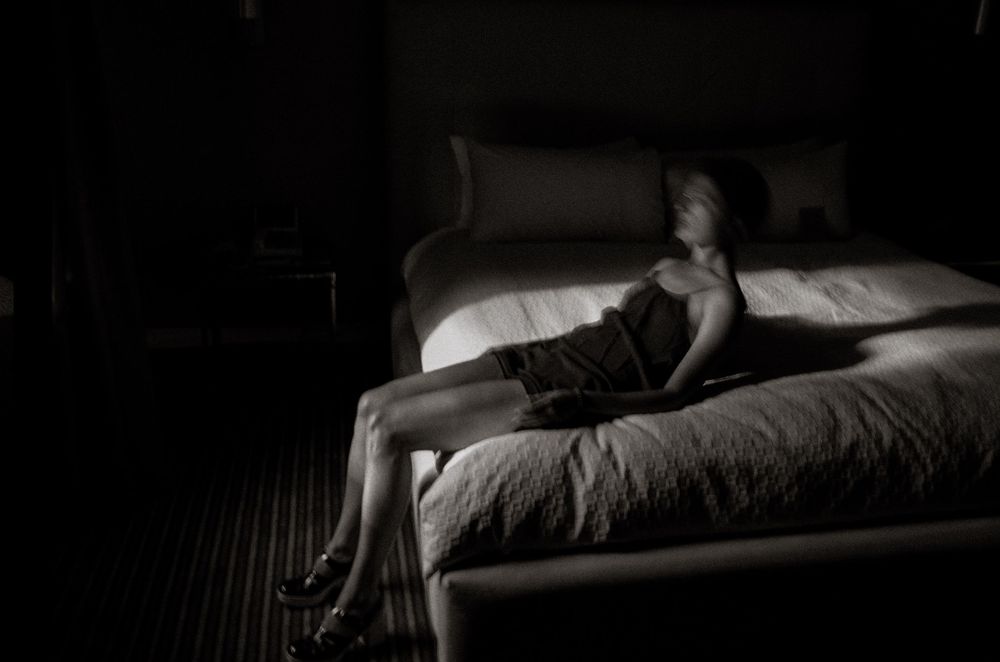 Woman in dark dress sits alone on white bed in dimly lit room, dramatic black and white photography.