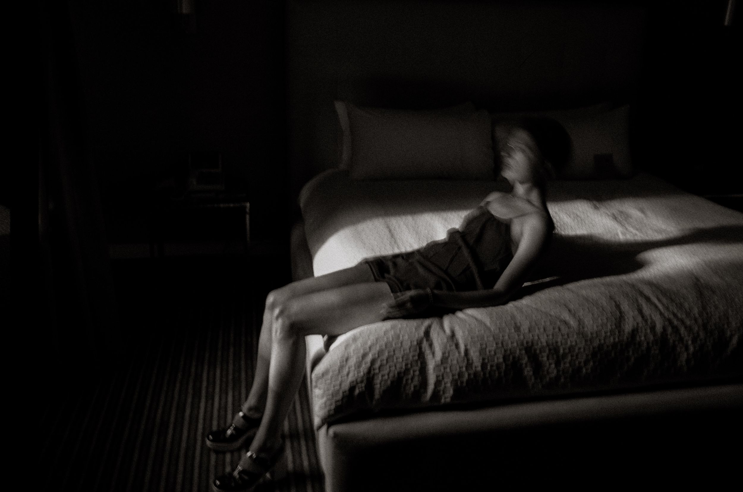 Woman in dark dress sits alone on white bed in dimly lit room, dramatic black and white photography.