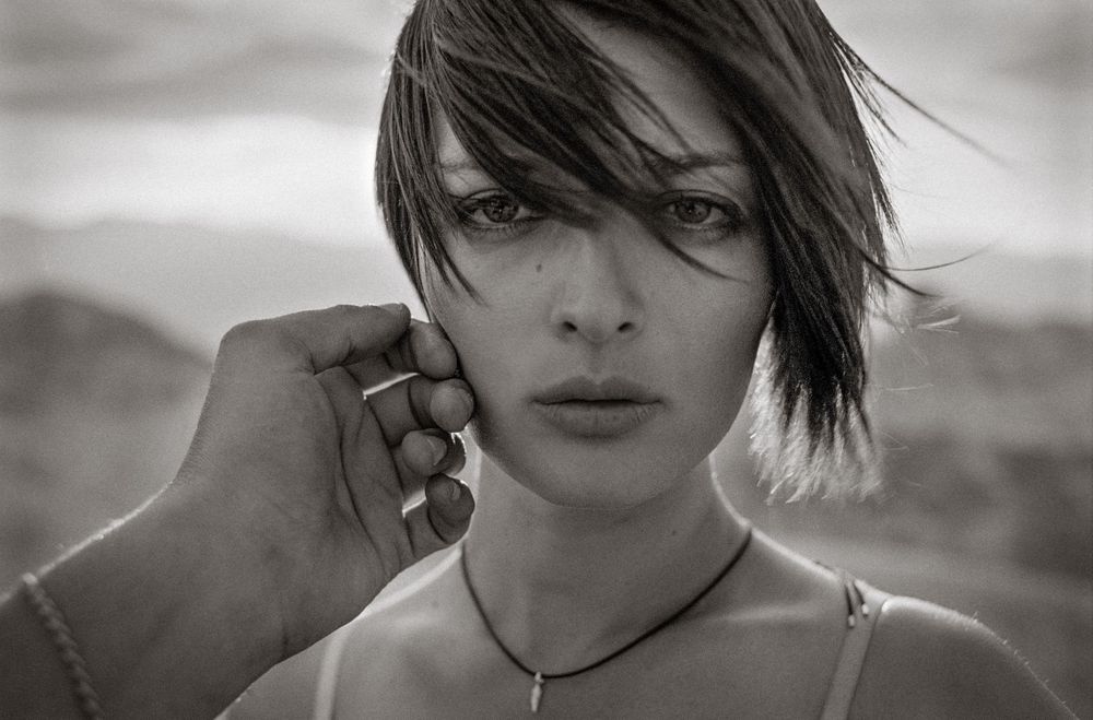 Black and white portrait of a young woman with short hair and a necklace, hand gently touching her face outdoors.