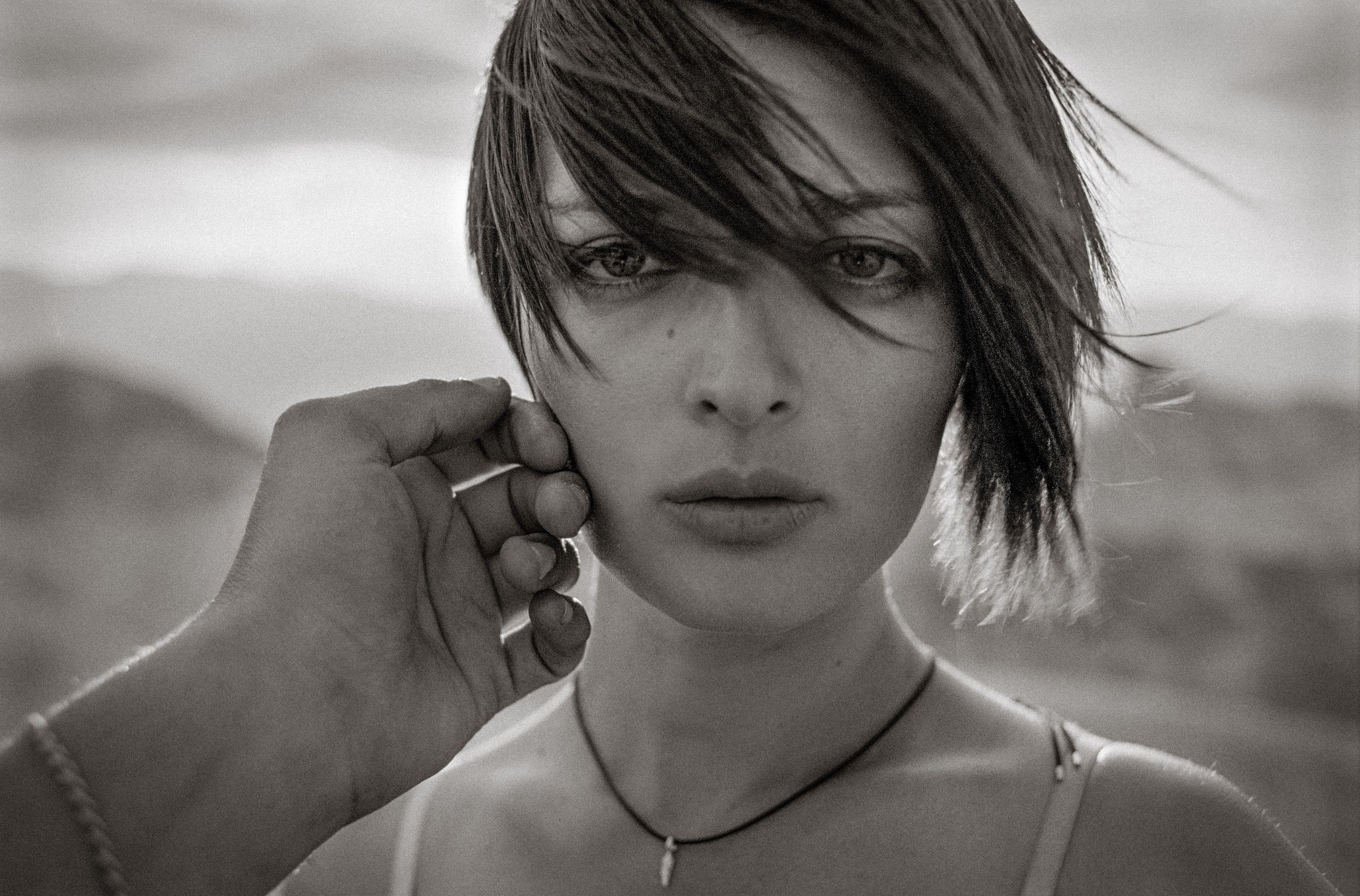 Black and white portrait of a young woman with short hair and a necklace, hand gently touching her face outdoors.