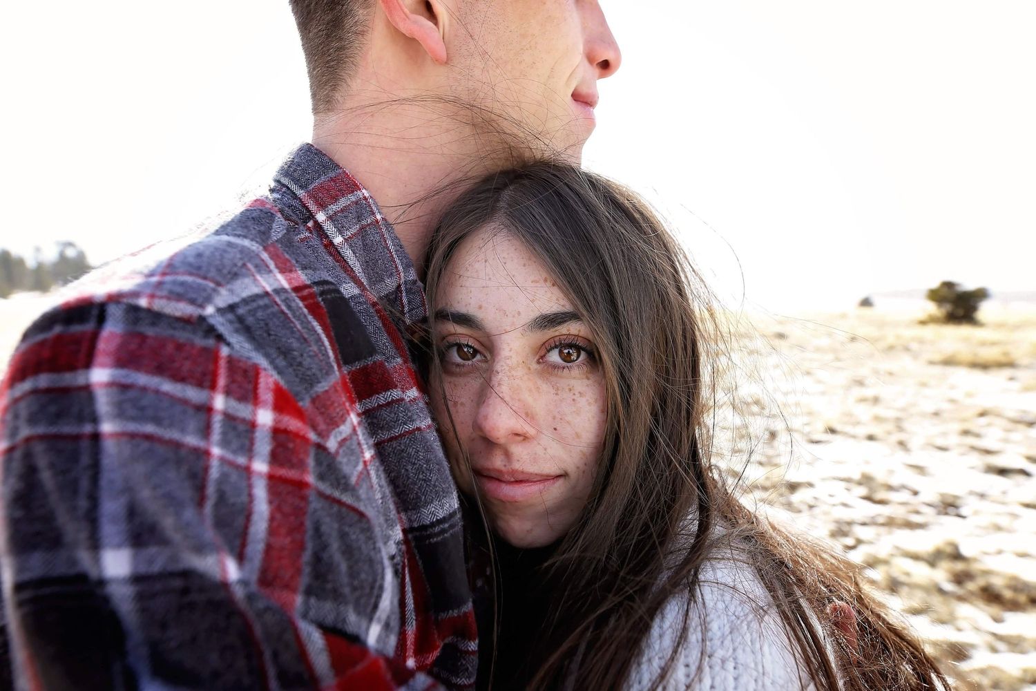 Young couple embraces outdoors in winter as a woman with long dark hair rests her head on a man in a plaid flannel shirt.