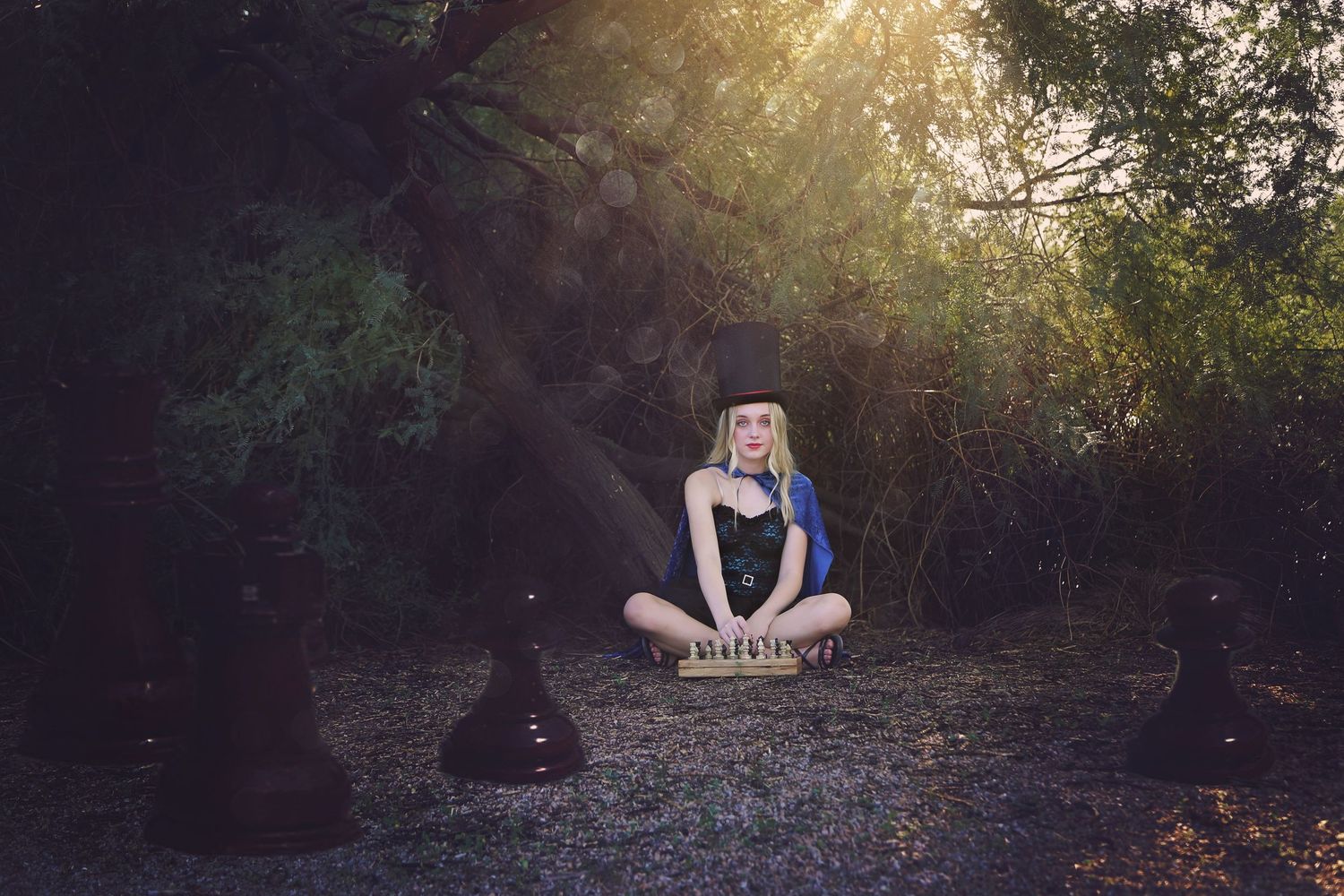Woman in a top hat sits cross-legged on a forest floor beside giant chess pieces as sunlight filters through the trees.