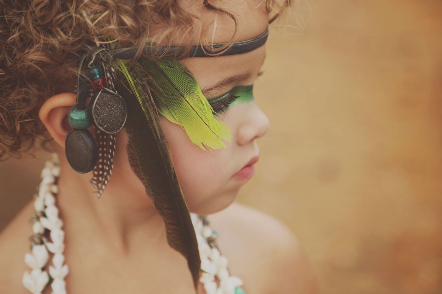 Profile of a young child with curly hair wearing a feather headband, green eye makeup, and a white shell necklace.