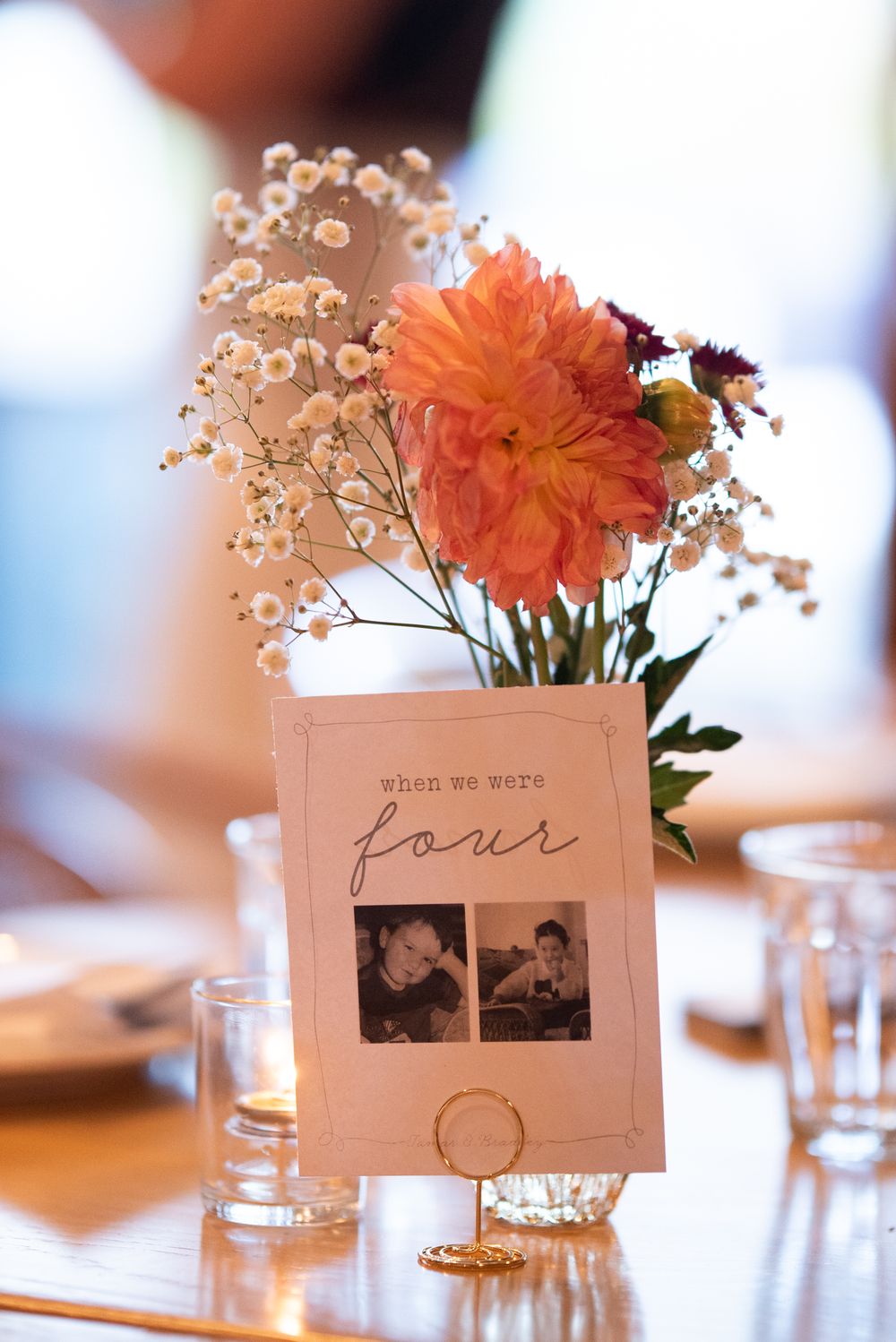 Wedding table centerpiece with orange dahlia, baby's breath, and a table number card reading 'when we were four' with photos.