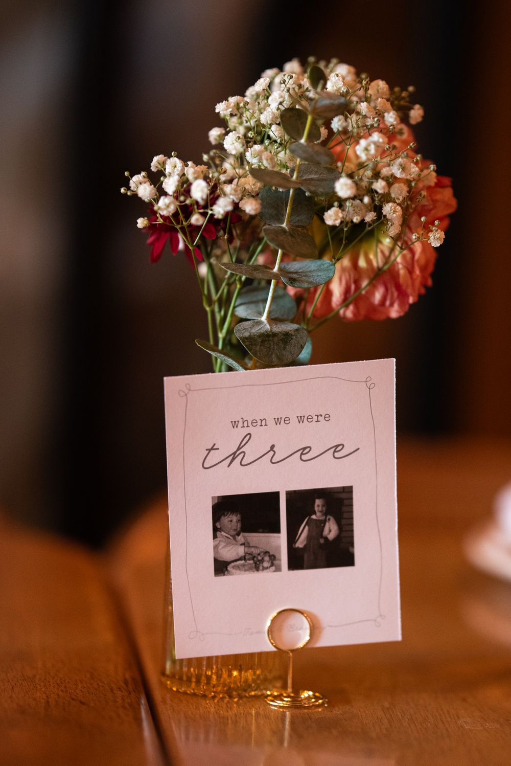 Wedding table number card reading 'when we were three' with black and white photos in gold holder beside floral arrangement.