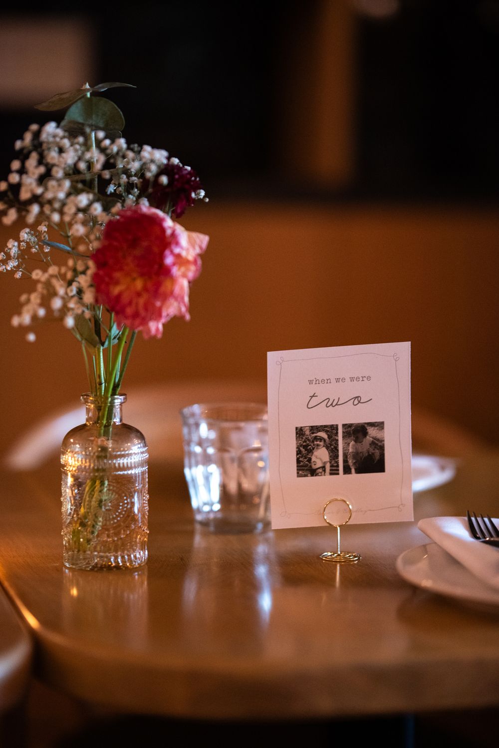 Elegant wedding table setting with pink dahlias in glass vase, table number card with photo, and gold ring holder.