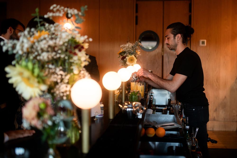 A bartender in a black shirt arranges glowing globe lights at a floral-decorated bar in a dimly lit venue.