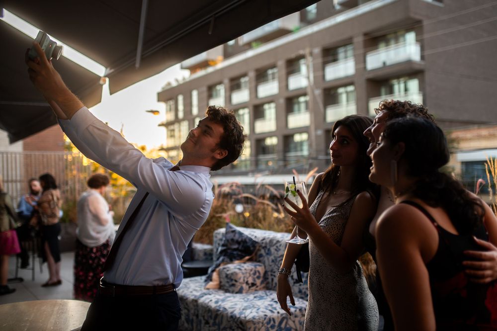 Group of friends taking a selfie at a rooftop party during sunset, holding drinks and smiling.