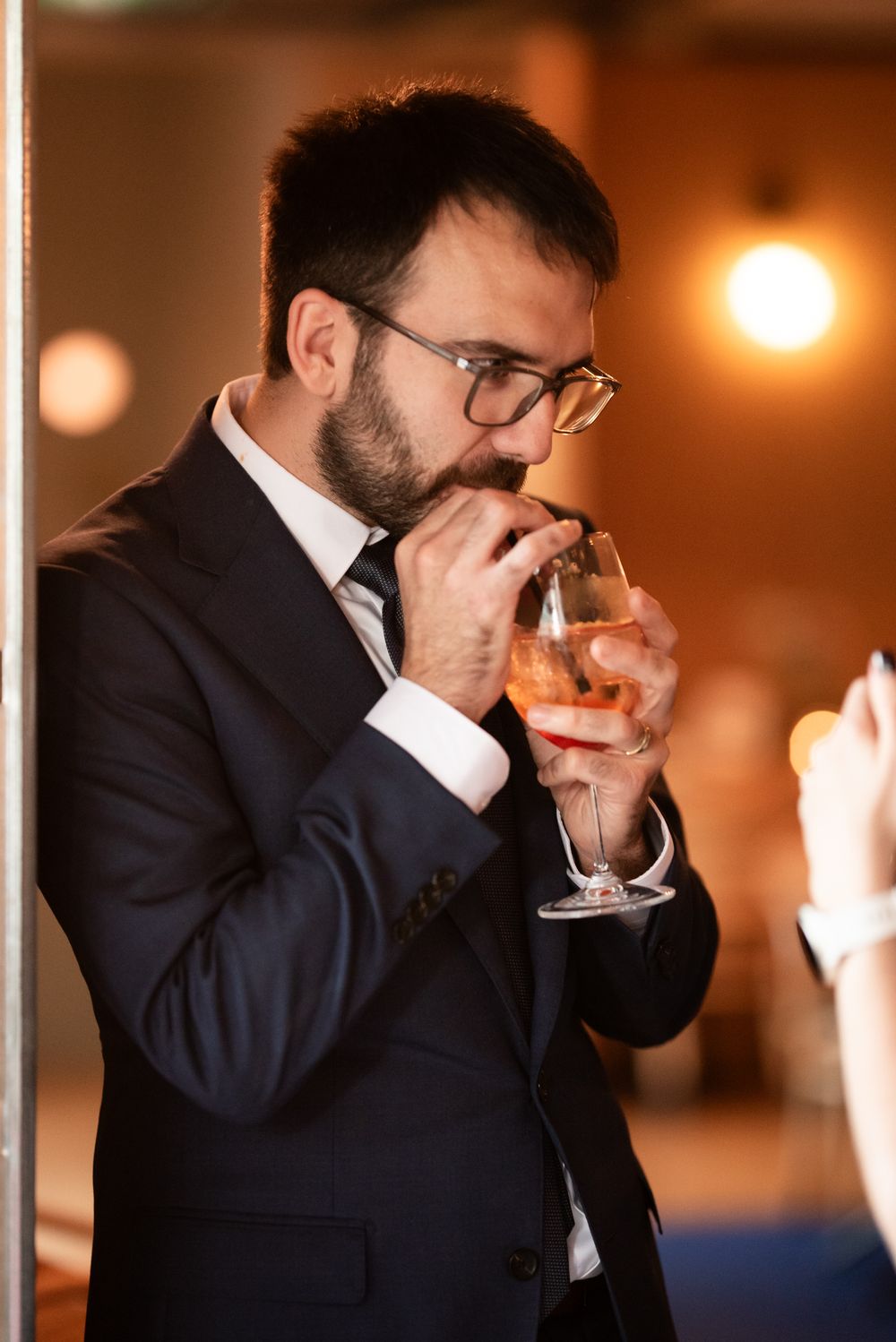 Bearded man in dark suit sipping rosé wine at an elegant indoor event or reception.