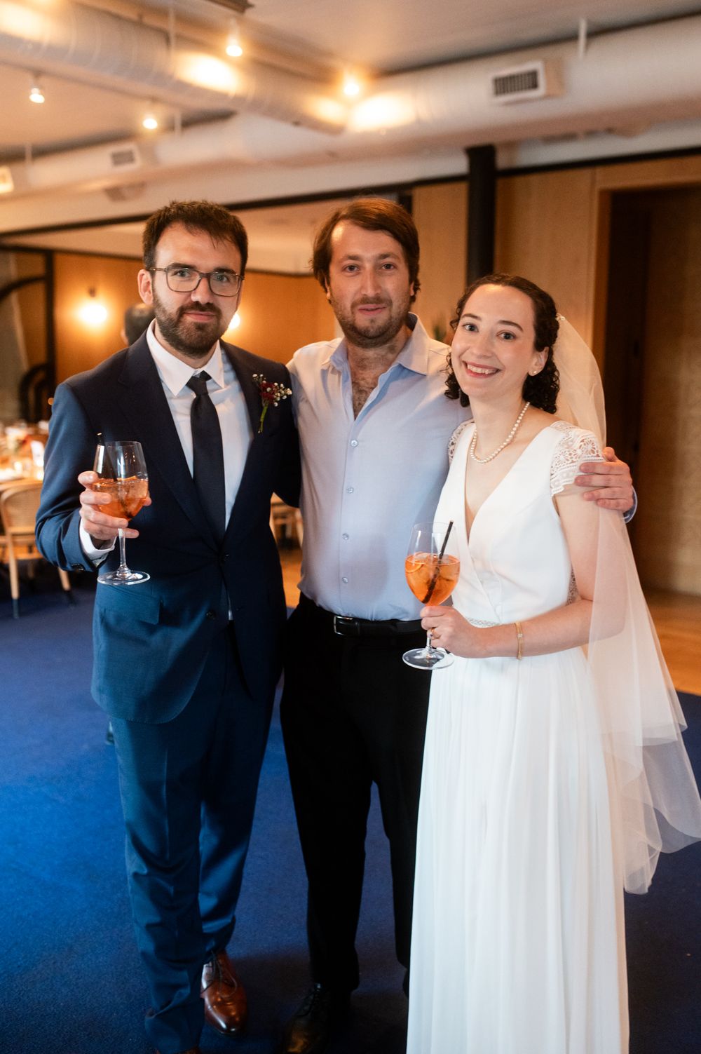 Bride in white gown and veil poses with groom in navy suit and guest holding Aperol spritz at wedding reception.