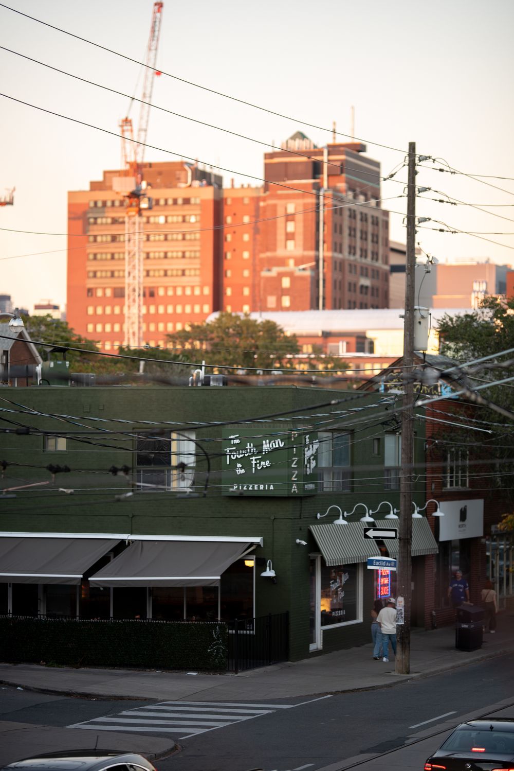 Urban street view of green pizzeria storefront with tall red brick building and cranes in background.