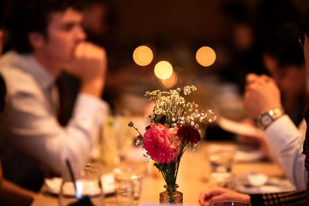 Elegant dinner party table with pink floral centerpiece and candles, guests socializing in soft bokeh background.