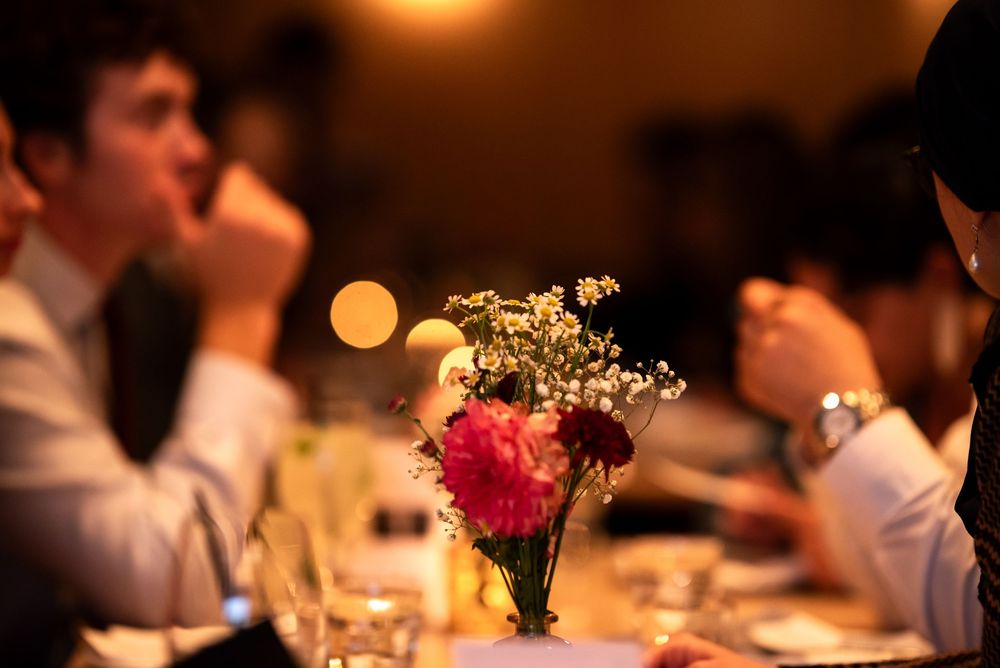 Elegant dinner party table with pink floral centerpiece and guests in formal attire in warm ambient lighting.