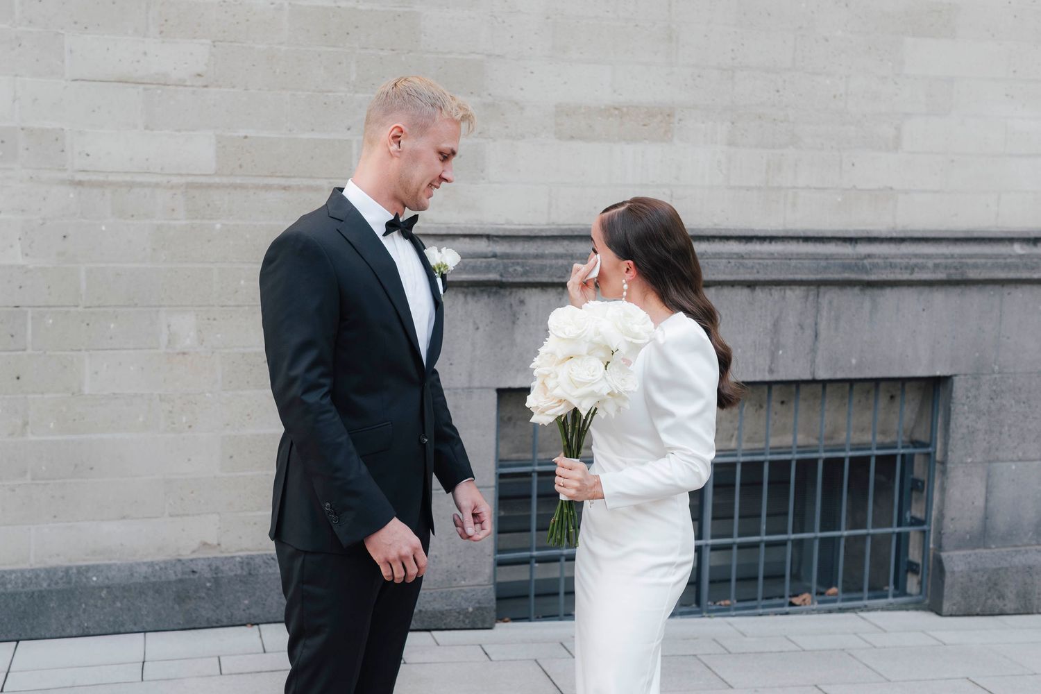 Bride in white gown holding white bouquet faces groom in black tuxedo outdoors against stone wall.