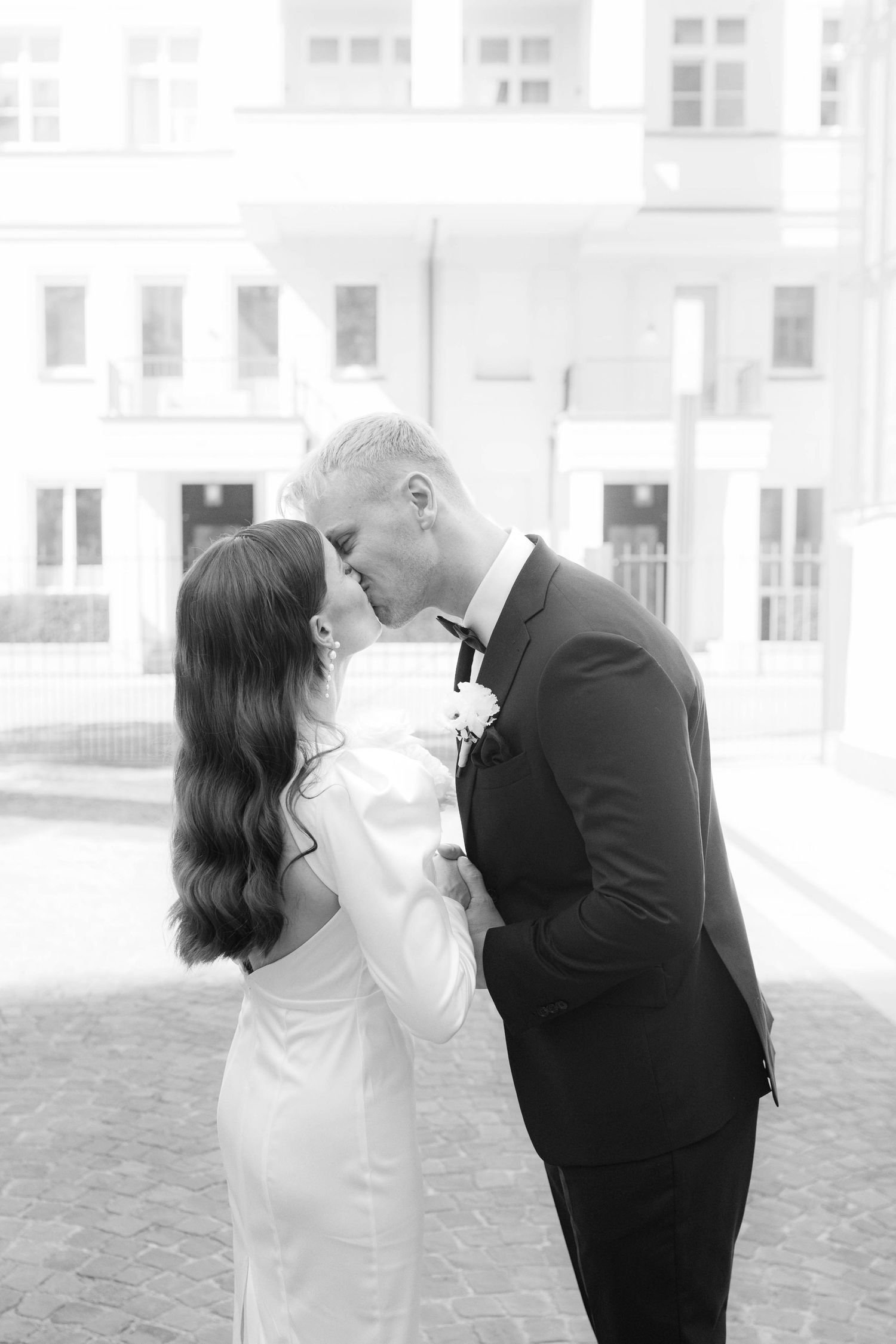 Black and white photo of a bride and groom sharing a kiss outdoors in front of a classic building.