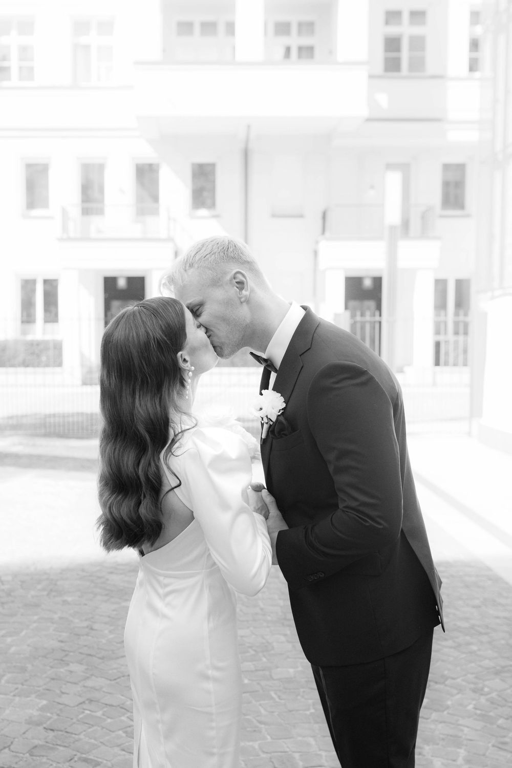 Black and white photo of a bride and groom sharing a kiss outdoors in front of a classic building.