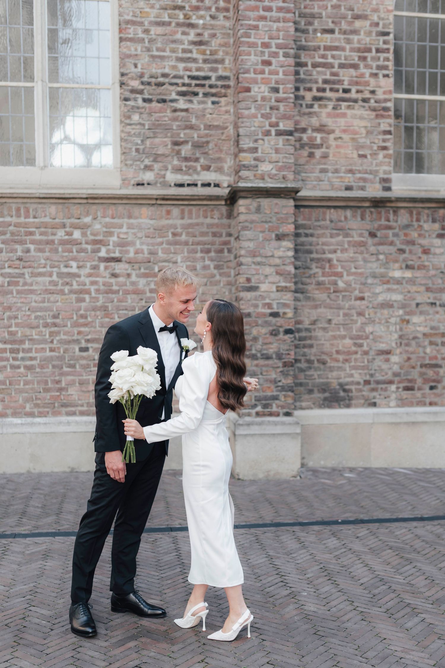 Bride and groom share a romantic moment outside a brick building, she in a white dress, he in a black tuxedo holding white flowers.