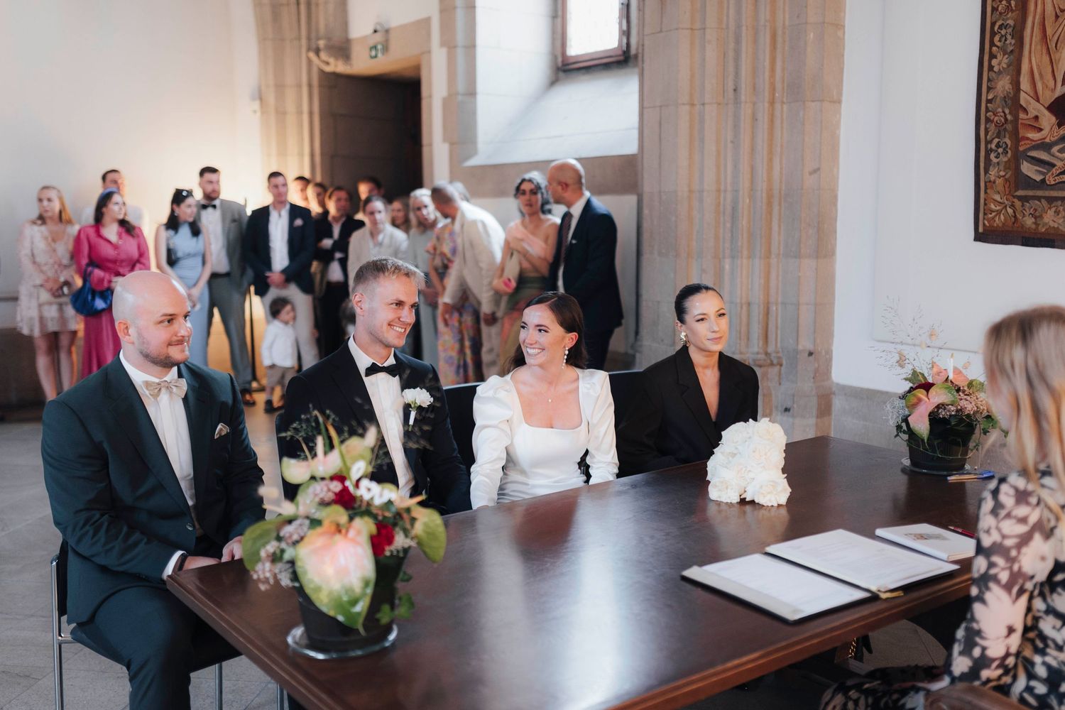 Elegant wedding ceremony with bride and groom seated at a table, guests gathered in a bright hall.