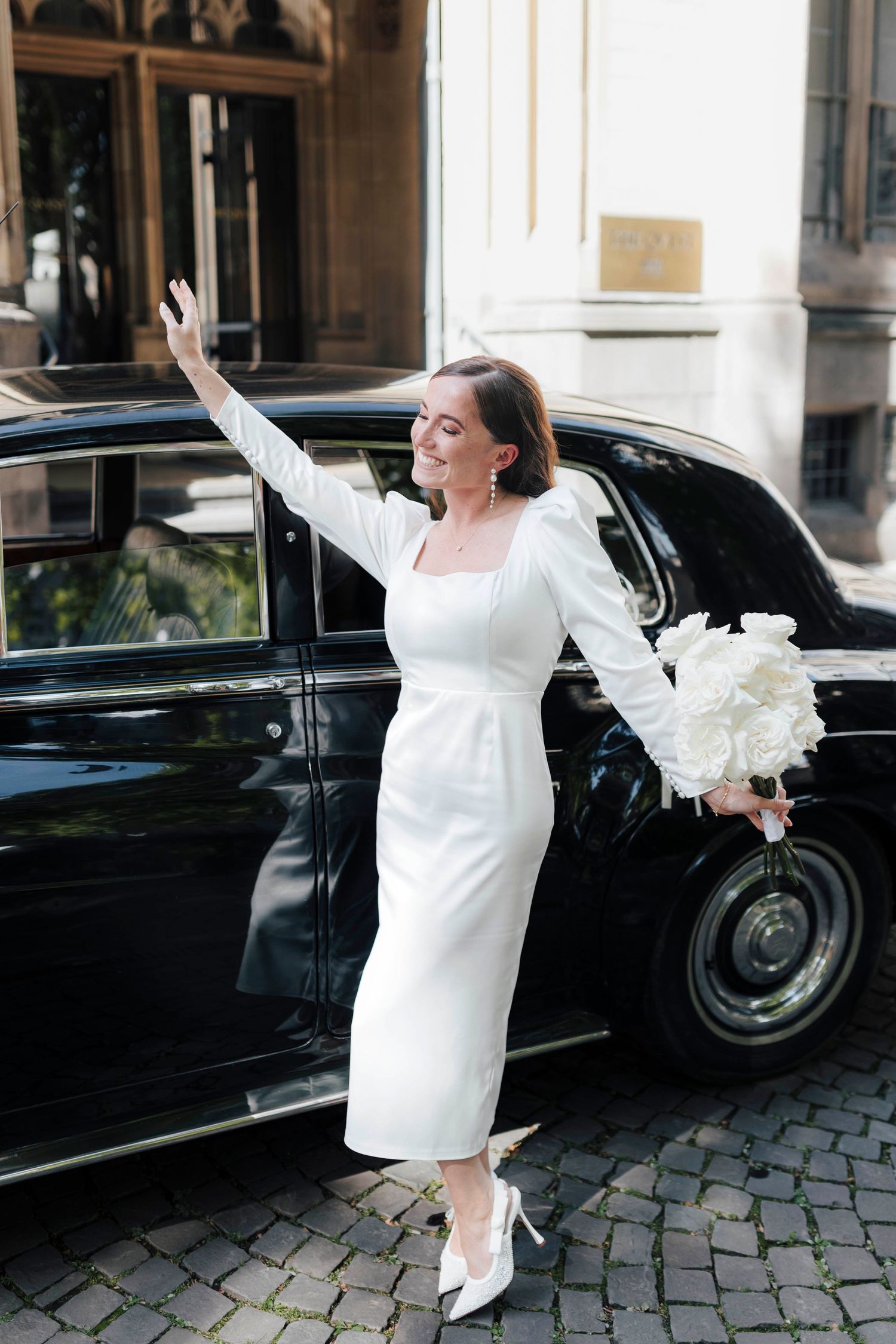 Elegant bride in white midi dress holding peony bouquet, waving beside a classic black vintage car on cobblestone street.