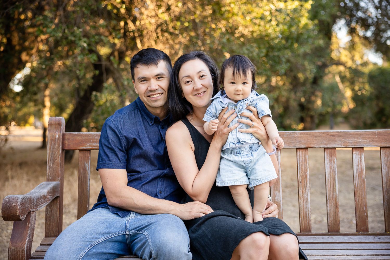 Happy family of three sitting on a wooden park bench, parents smiling with baby in outdoor Bay Area setting.