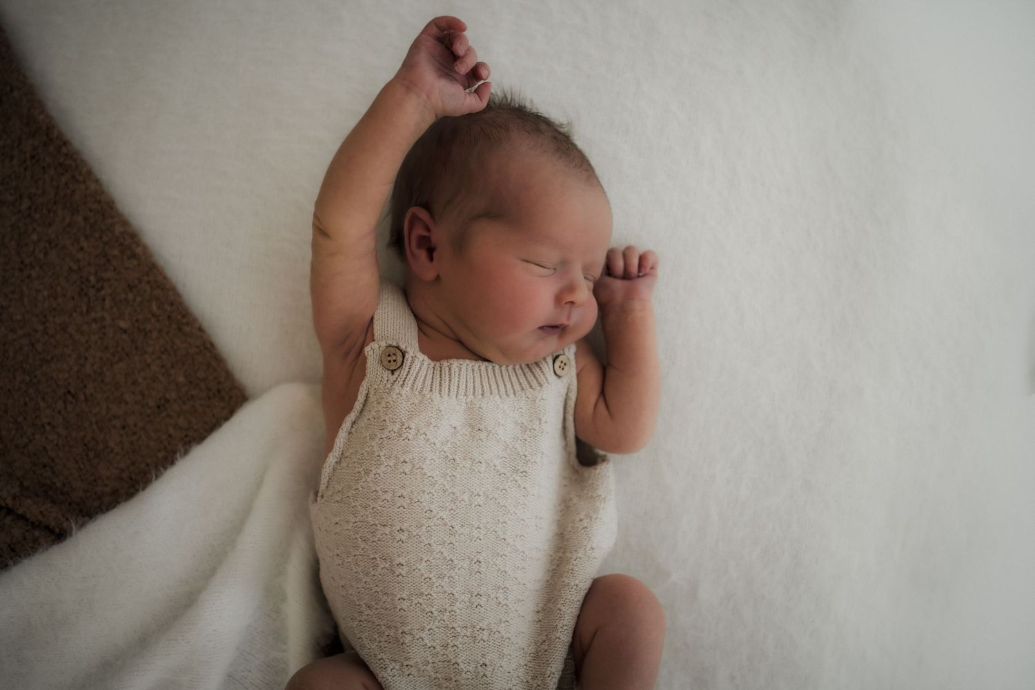 Sleeping newborn baby in cream knit romper lying on white blanket with one arm raised.