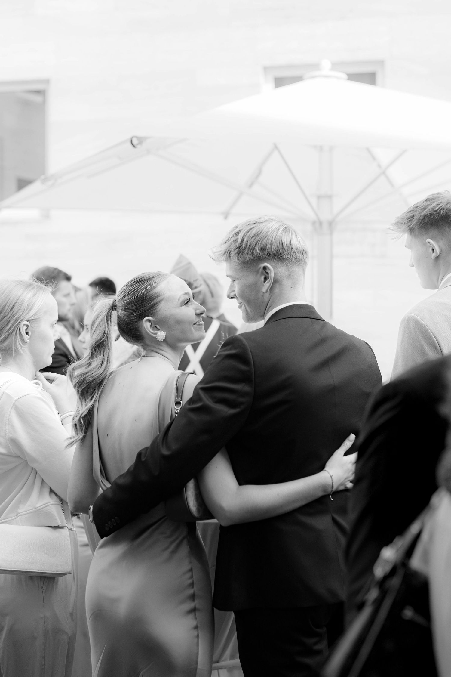 Black and white photo of elegantly dressed guests embracing and socializing at an outdoor wedding reception.