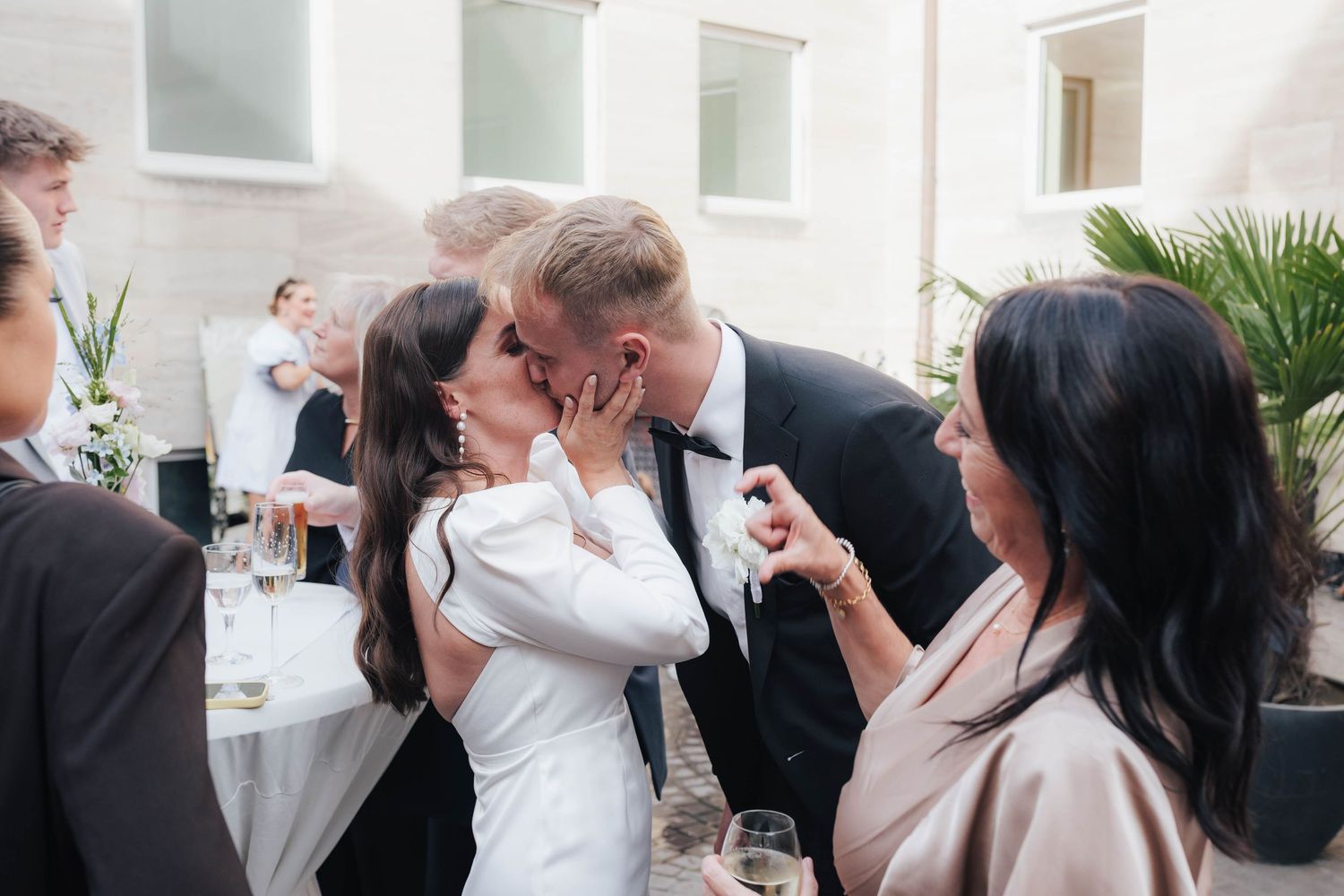 Bride and groom share a kiss at their wedding reception surrounded by guests in elegant attire.