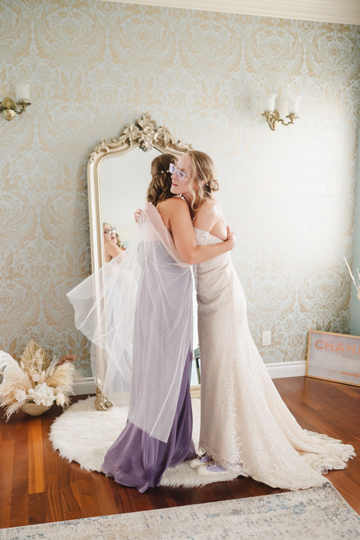Two brides in elegant wedding gowns share a tender embrace in front of an ornate gold mirror in a beautifully decorated room.