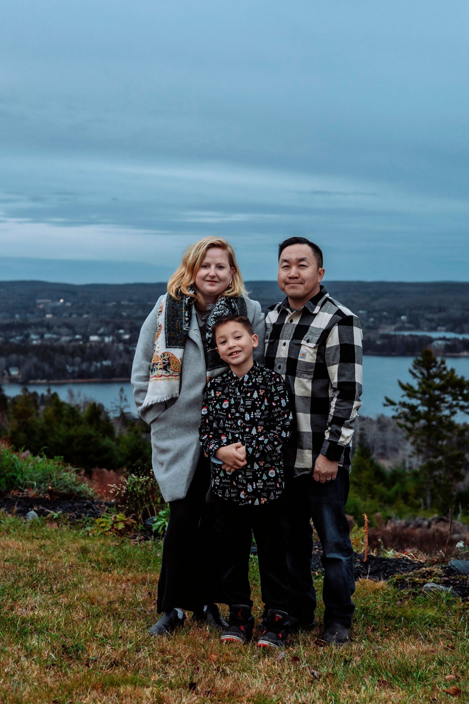 Family of three posing on a hilltop with a scenic coastal town and cloudy blue sky in the background.