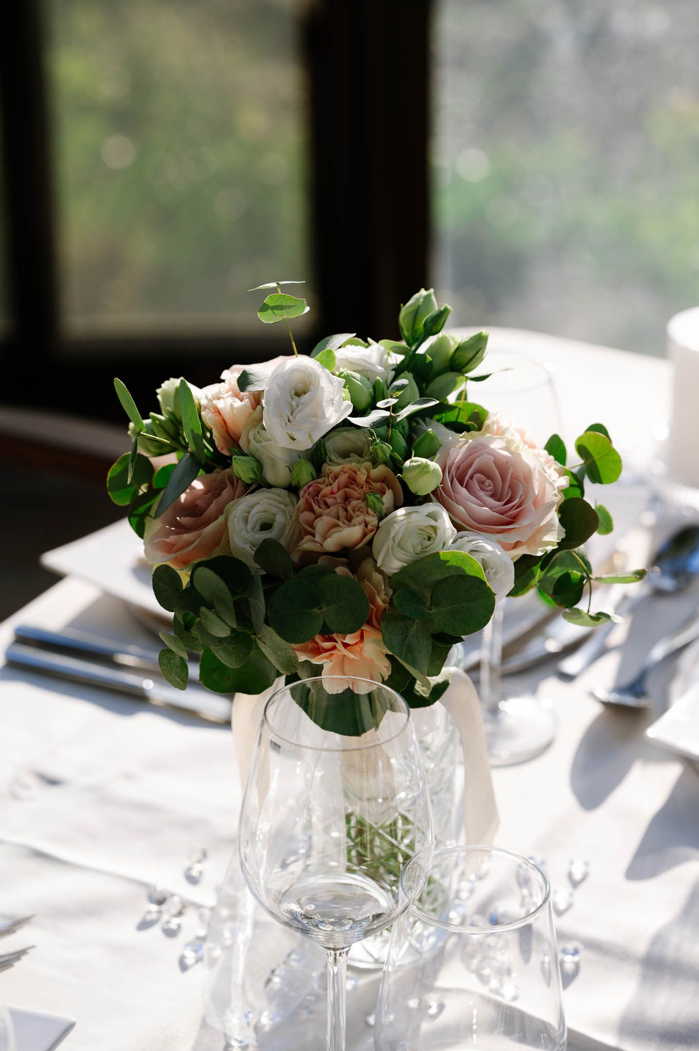 Elegant wedding table centerpiece featuring pink and white roses with lush green foliage in glass vase.