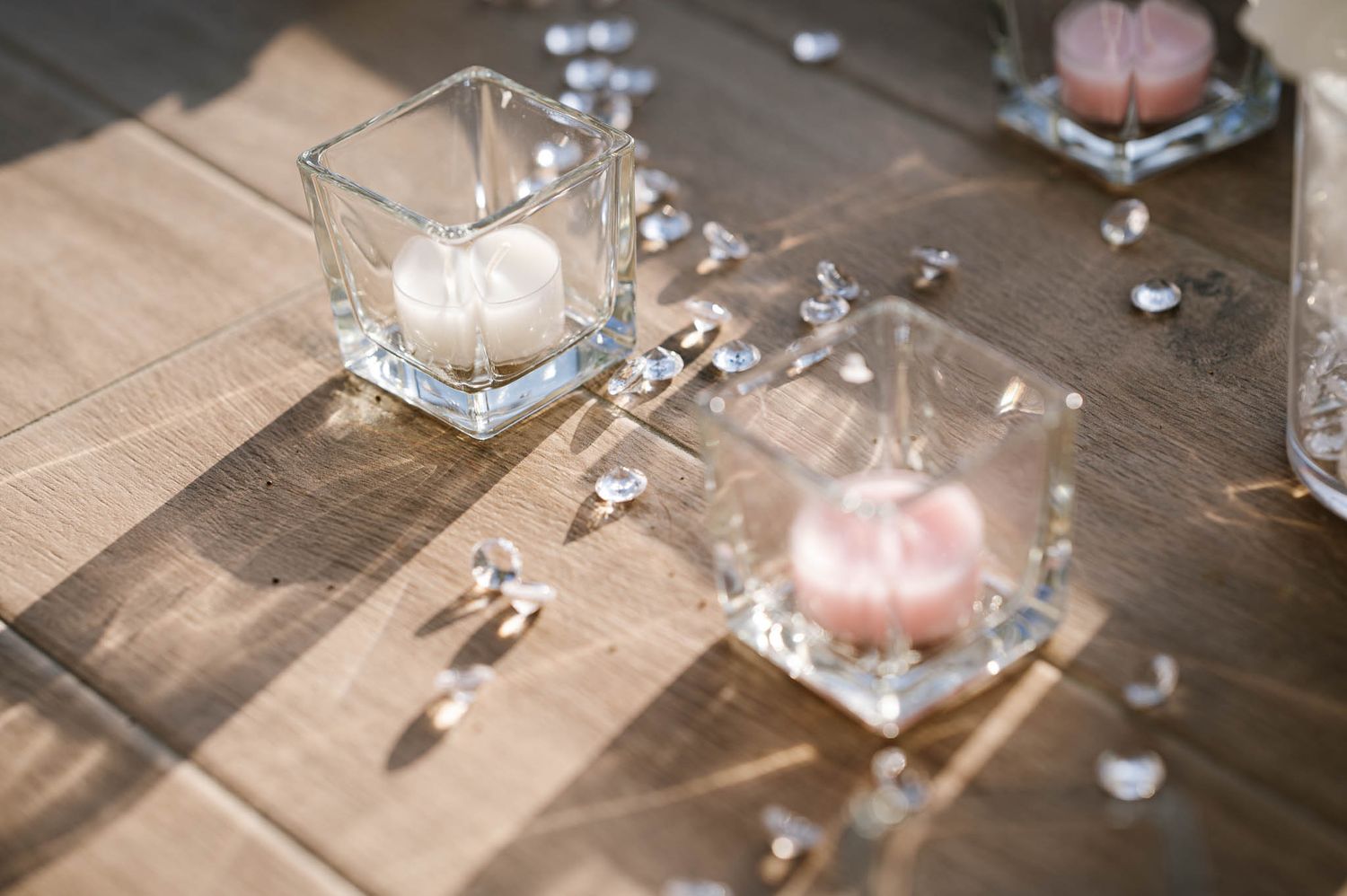 Elegant wedding table decor featuring square glass candle holders with white and pink candles scattered with crystal gems.