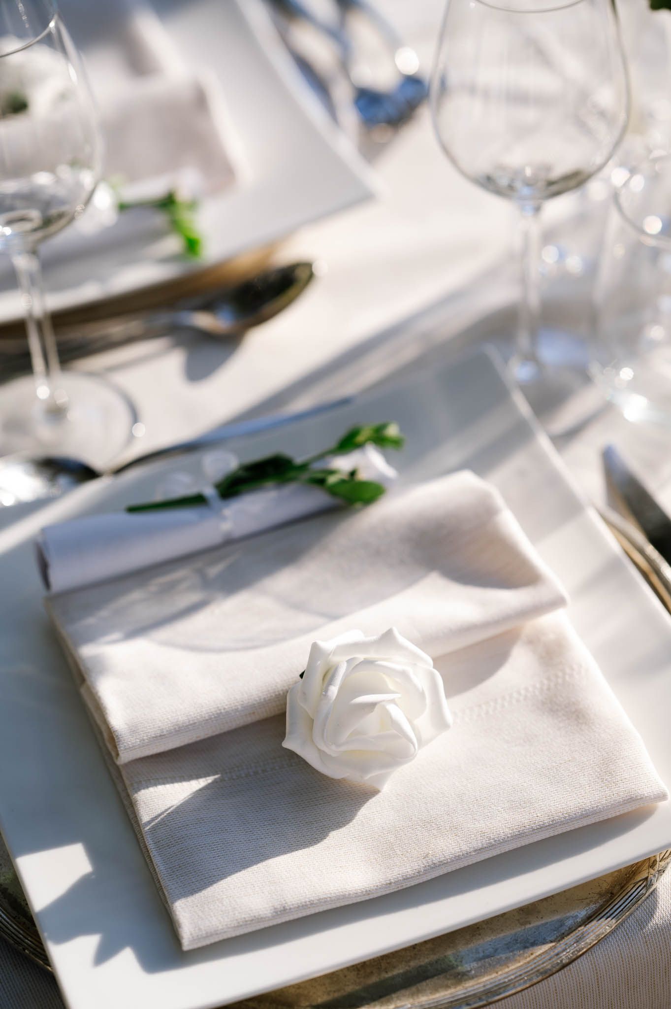 Elegant white wedding place setting with folded napkin, white rose detail, and crystal wine glasses.