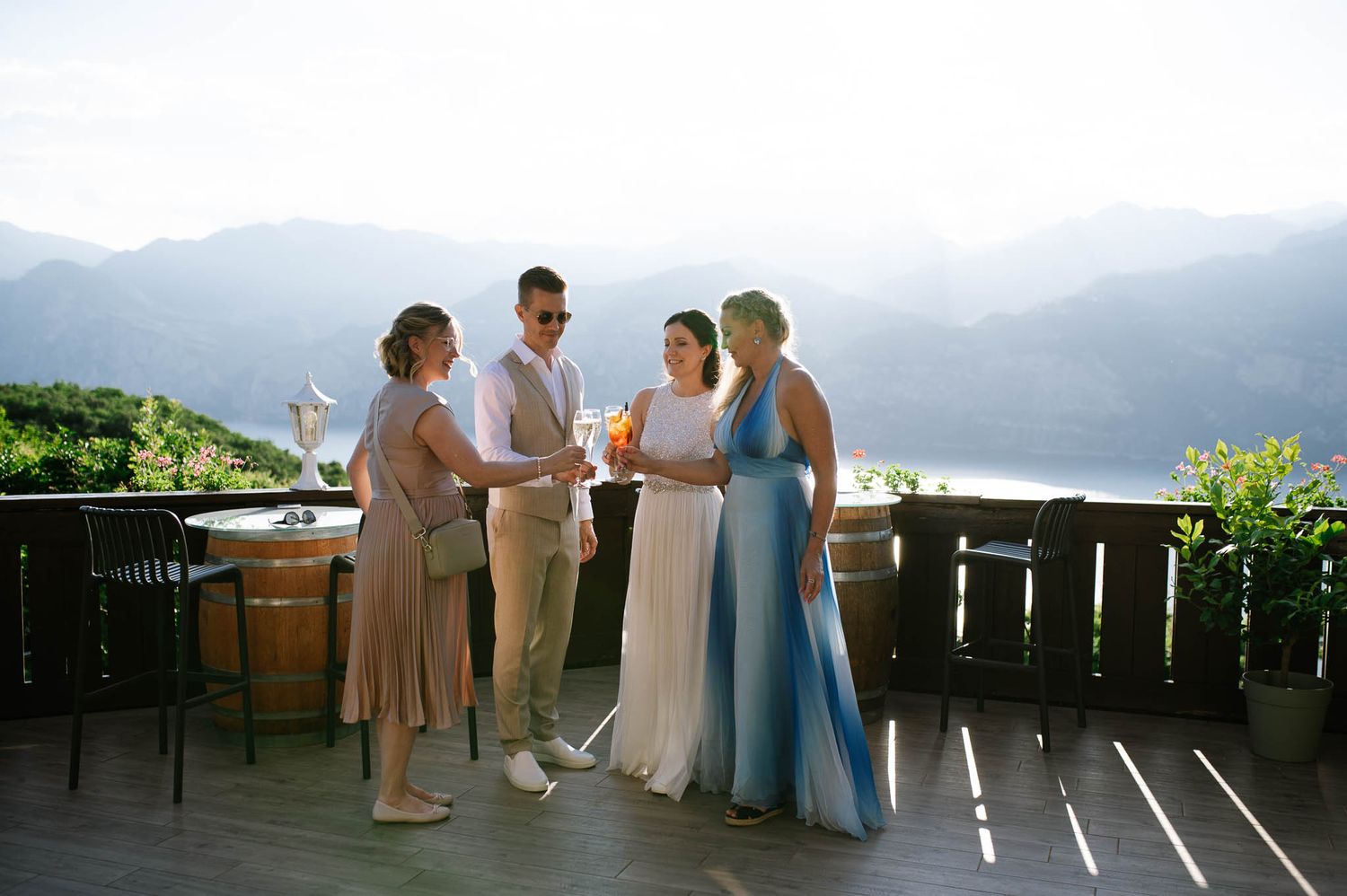 Wedding party socializing on a wooden terrace with breathtaking mountain and lake views in background.