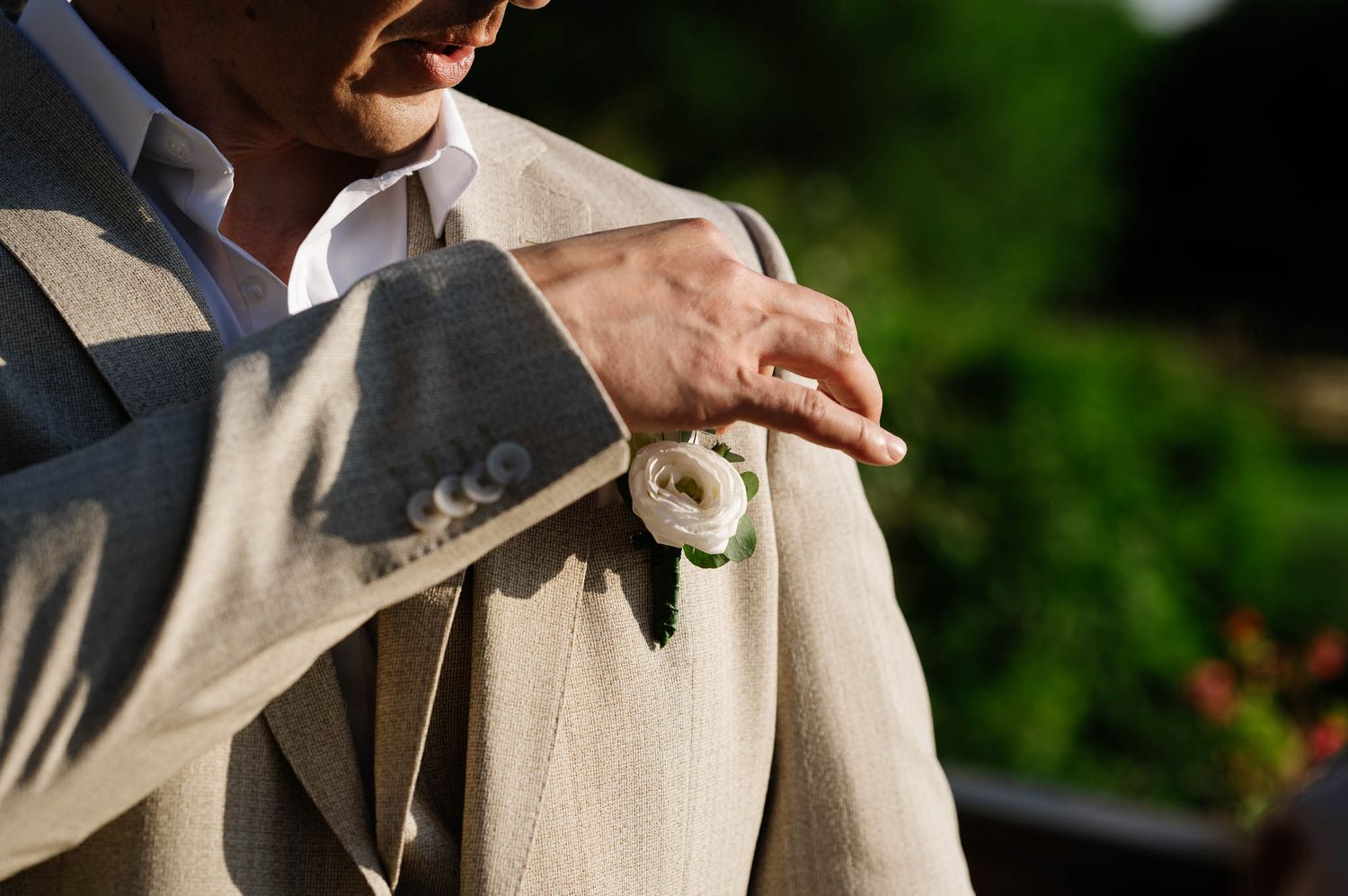 Groom in beige suit adjusts white rose boutonniere on lapel outdoors.