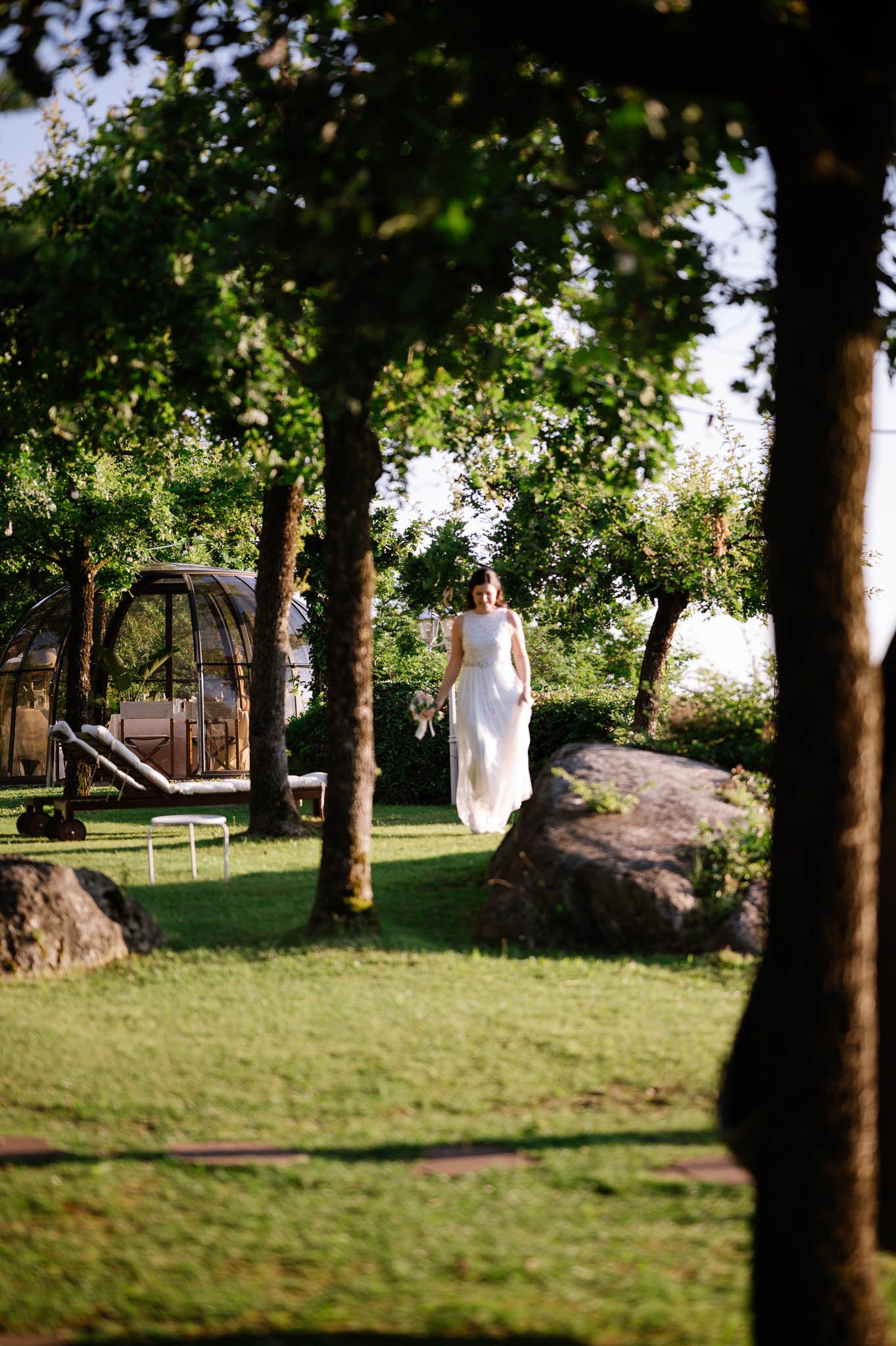 Bride in white gown walking through sunlit garden with lush green trees and lawn.