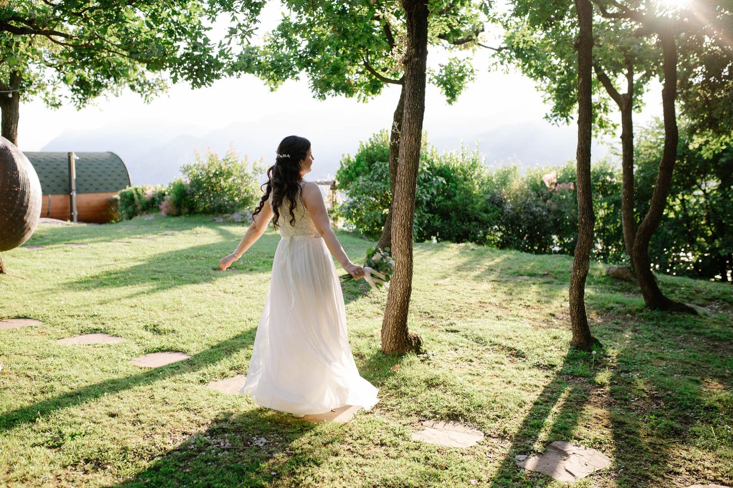Bride in flowing white gown walks through sunlit garden with lake views and lush green trees.