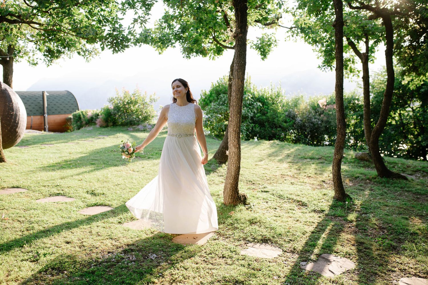 Bride in white gown walks through sunlit garden with trees and scenic countryside in background.