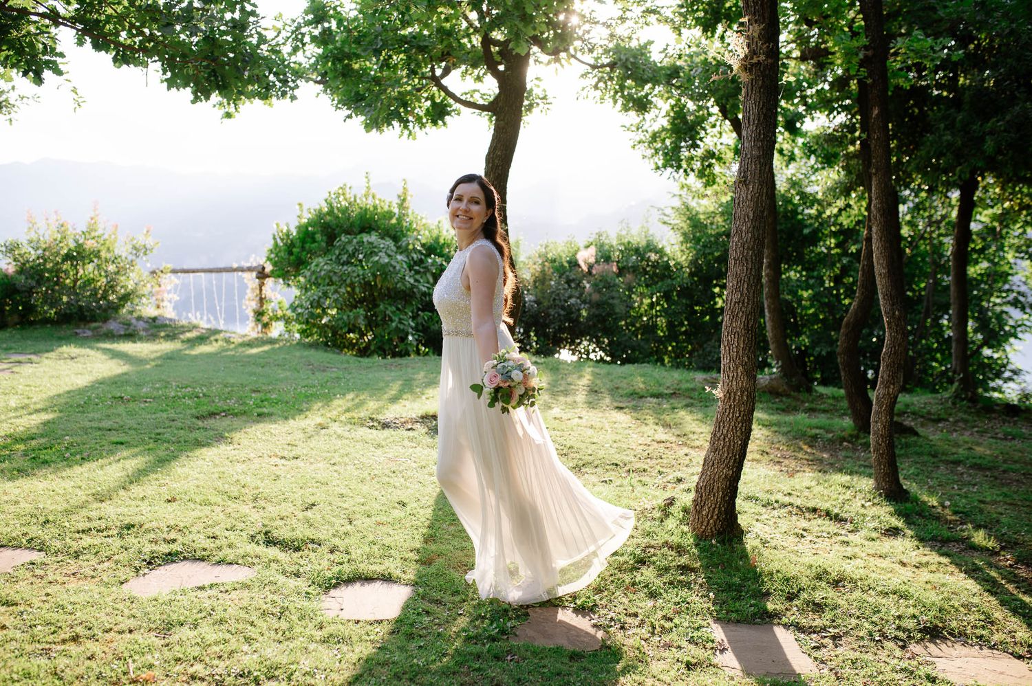 Elegant bride holding bouquet strolls through sun-drenched garden with stunning lake backdrop.
